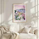 a framed poster of a garden scene with flowers and a lighthouse, hanging on a wall above a white couch with pillows.