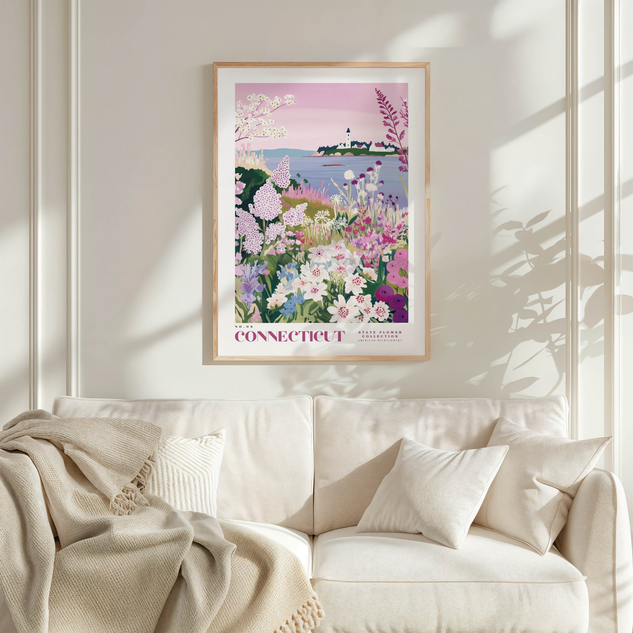 a framed poster of a garden scene with flowers and a lighthouse, hanging on a wall above a white couch with pillows.