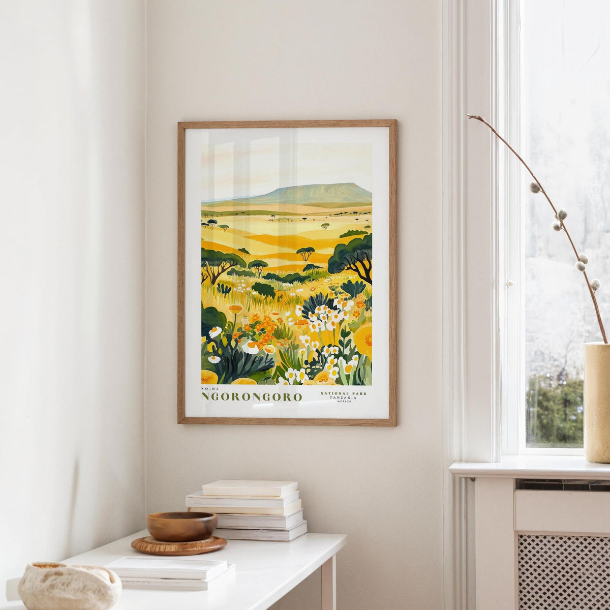 a framed poster of a landscape with yellow flowers hanging on a wall in a room with a white table and a window.