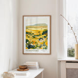 a framed poster of a landscape with yellow flowers hanging on a wall in a room with a white table and a window.