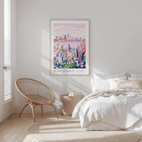 A bedroom with a large framed poster of a cityscape of Wisconsin hanging on the wall, featuring a mix of pink, purple, and green colors. The poster is positioned above a bed with white bedding and pillows. A woven chair is also present in the room.