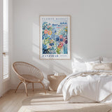 a bedroom with a bed, a woven chair, and a framed poster of a colorful floral scene hanging on the wall.
