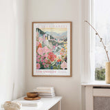 a framed poster of a cityscape with flowers hanging on a wall, next to a window and a small table with books and a bowl.