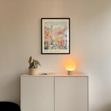 a white cabinet with a potted plant and a lamp on top, set against a wall with a framed poster of a flower market in San Antonio.