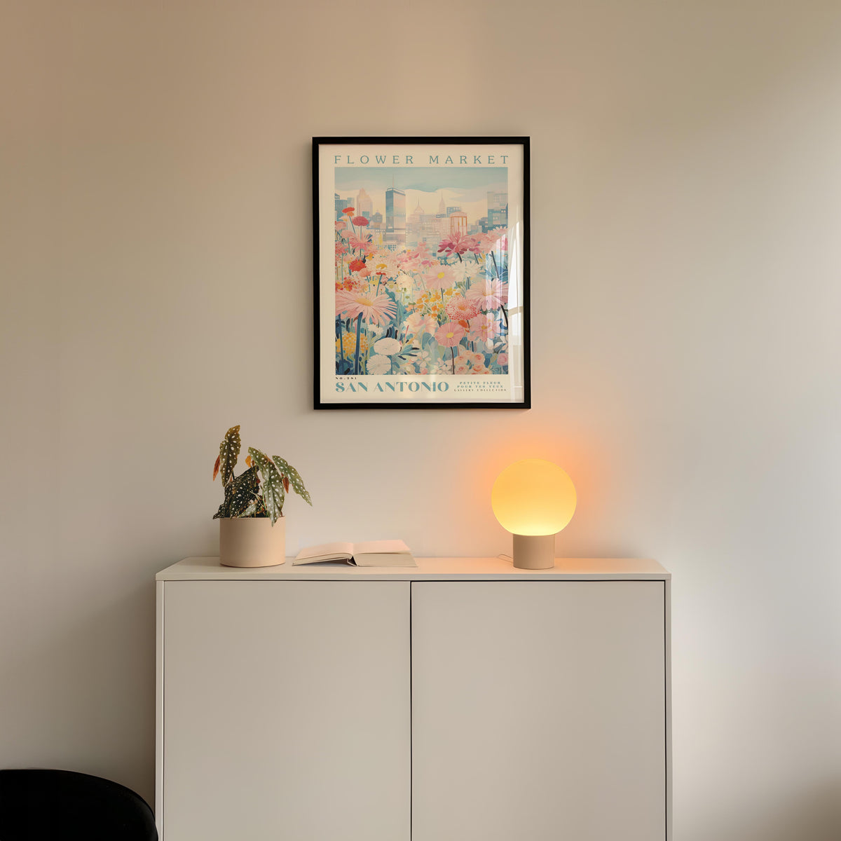 a white cabinet with a potted plant and a lamp on top, set against a wall with a framed poster of a flower market in San Antonio.