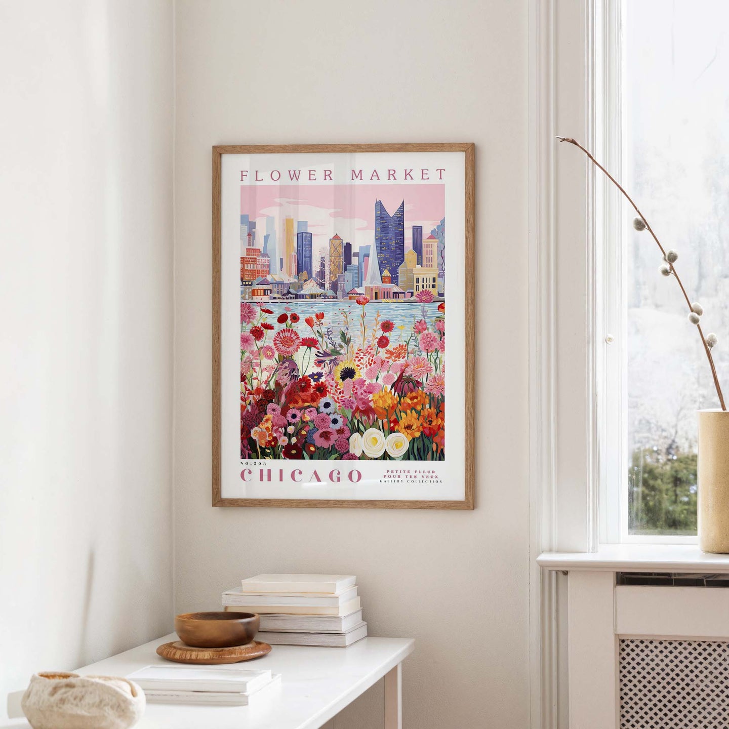 a framed poster of a cityscape with a flower market in Chicago, hanging on a wall in a room with a window and a small table with books and a vase.