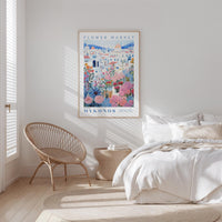 a bedroom with a bed, a woven chair, and a framed poster of a flower market on the wall.
