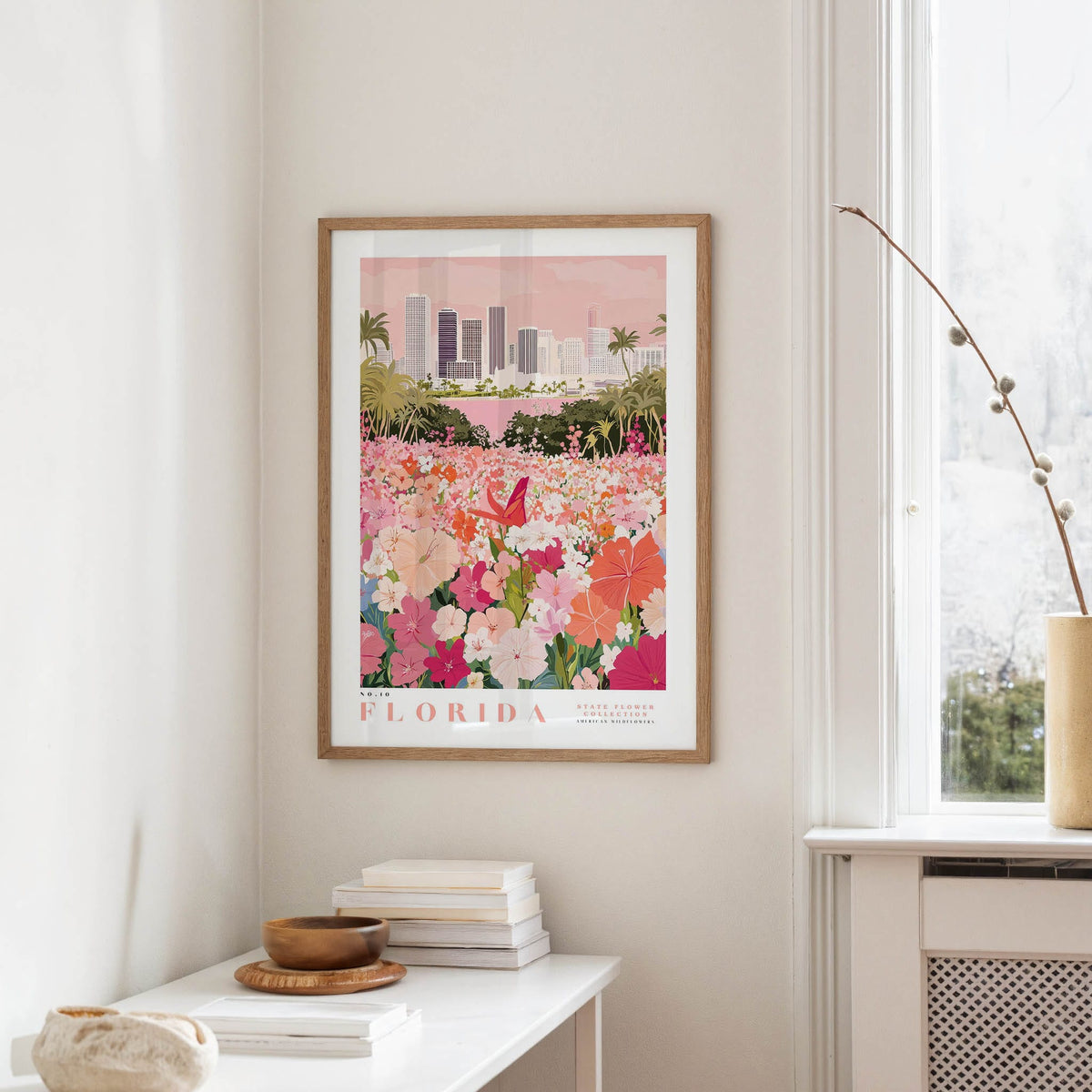 a framed poster of a colorful floral scene hanging on a wall, with a window and a small table with books and a vase nearby.