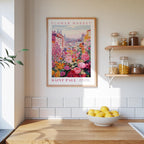 a kitchen scene with a framed poster of a colorful flower market in Saint Paul, Minnesota, hanging on the wall above a wooden counter. The poster is surrounded by various kitchen items, such as jars, a bowl of lemons, and a plant.