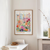 a framed poster of a colorful floral scene hanging on a wall, with a white table and a vase of flowers in the foreground.
