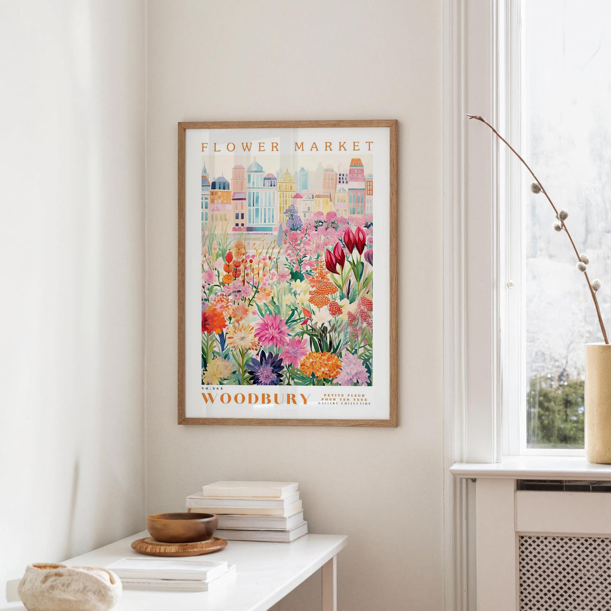a framed poster of a colorful floral scene hanging on a wall, with a white table and a vase of flowers in the foreground.