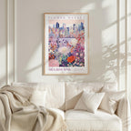a framed poster of a cityscape with a flower market in the background, hanging on a wall above a white couch with a blanket draped over it.
