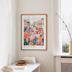 a framed poster of a colorful floral scene hanging on a wall, with a white table and a vase of flowers in the foreground.