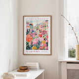 a framed poster of a colorful floral scene hanging on a wall, with a white table and a vase of flowers in the foreground.