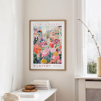 a framed poster of a colorful floral scene hanging on a wall, with a white table and a vase of flowers in the foreground.