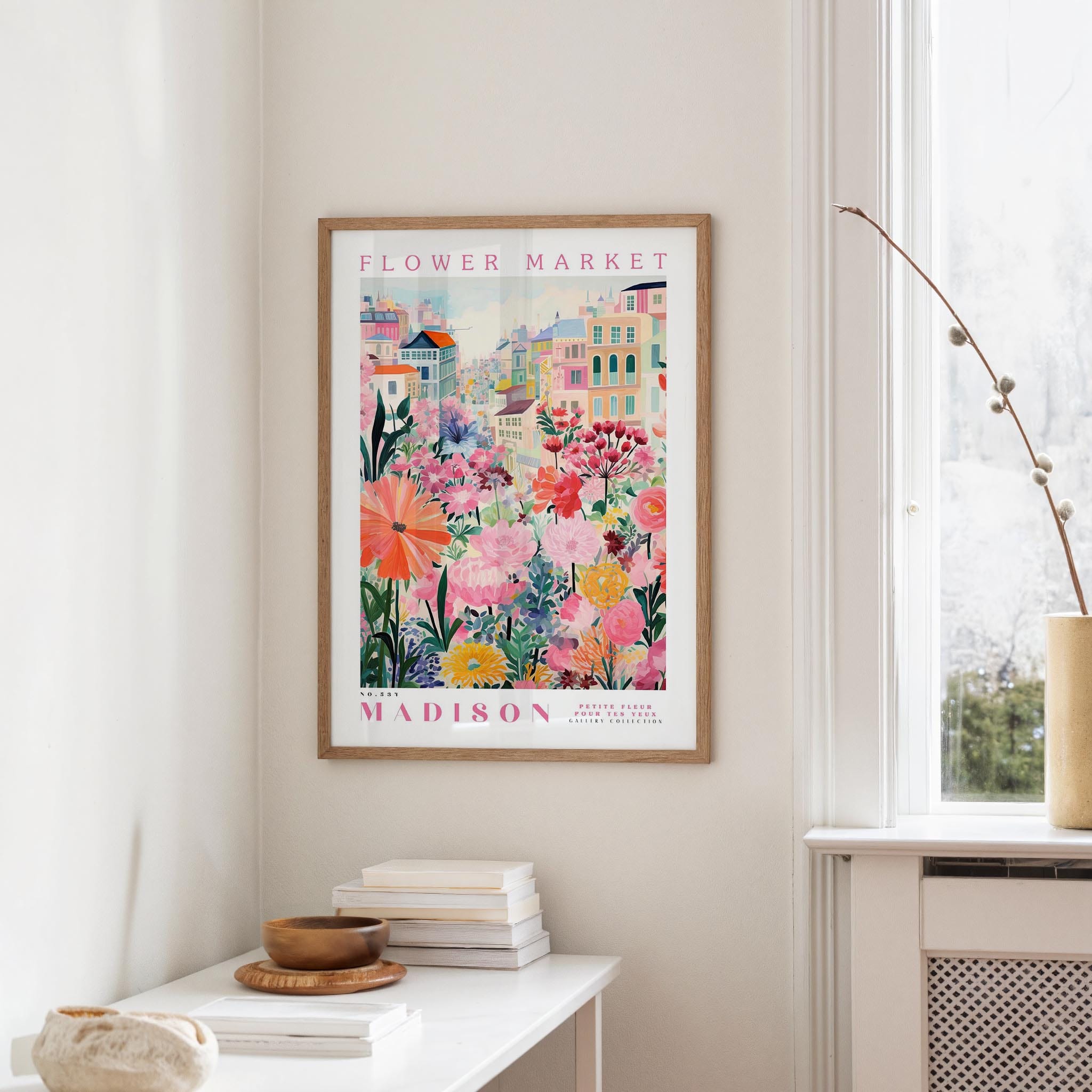 a framed poster of a colorful floral scene hanging on a wall, with a white table and a vase of flowers in the foreground.