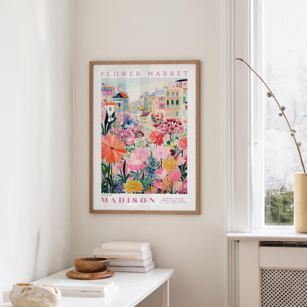 a framed poster of a colorful floral scene hanging on a wall, with a white table and a vase of flowers in the foreground.