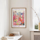 a framed poster of a colorful floral scene hanging on a wall, with a white table and a vase of flowers in the foreground.