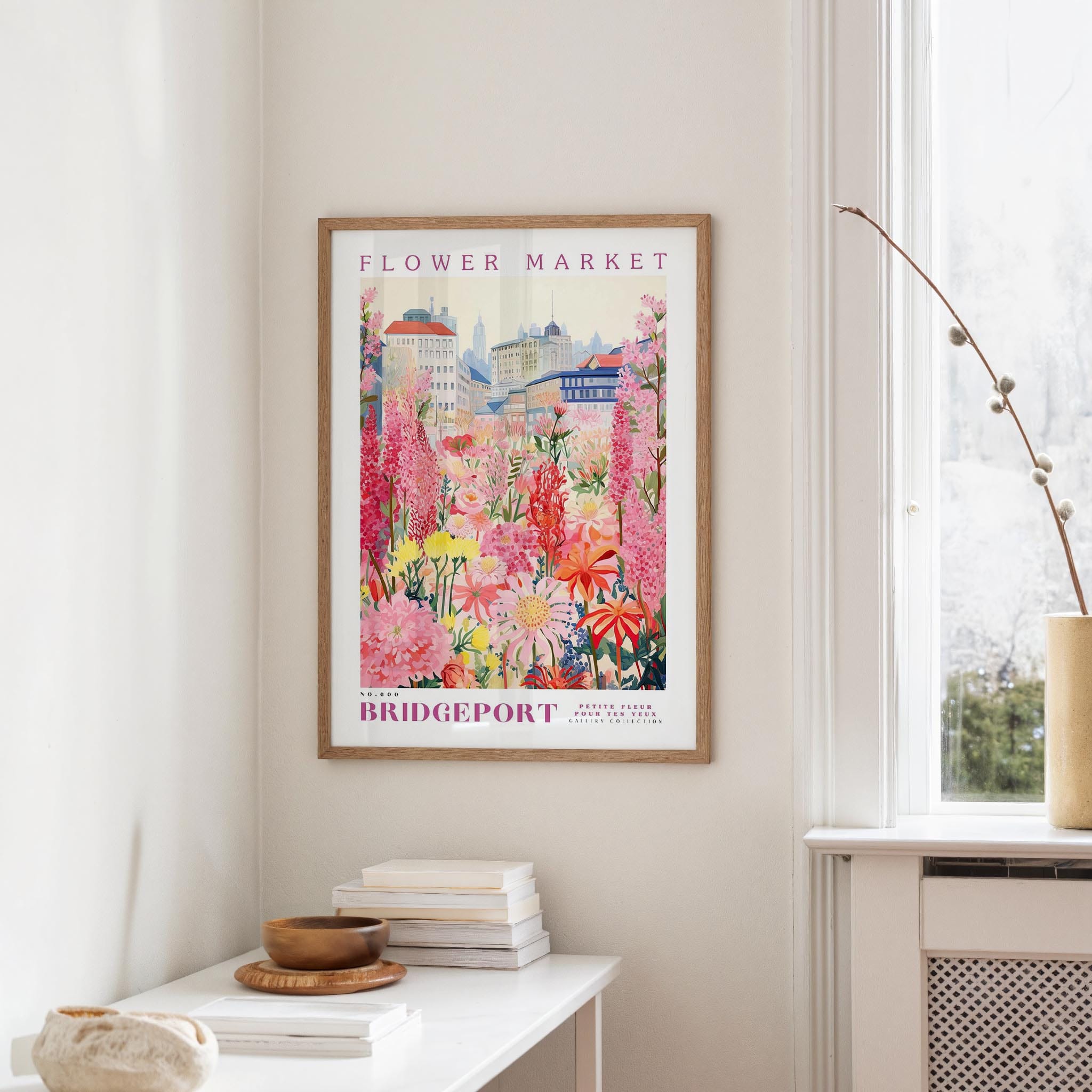a framed poster of a colorful floral scene hanging on a wall, with a white table and a vase of flowers in the foreground.