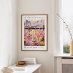 a framed poster of a cityscape with a field of colorful flowers in the foreground, hanging on a wall in a room with a window and a small table with books and a vase.