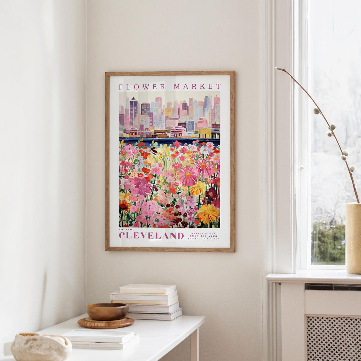 a framed poster of a cityscape with a field of colorful flowers in the foreground, hanging on a wall in a room with a window and a small table with books and a vase.