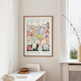 a framed poster of a flower market in Warsaw hanging on a wall, with a white table and a vase of flowers in the foreground.
