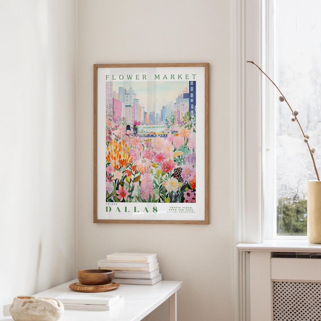 a framed poster of a colorful floral scene hanging on a wall, with a white table and a vase of flowers in the foreground.