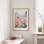 a framed poster of a colorful floral scene hanging on a wall, with a white table and a vase of flowers in the foreground.