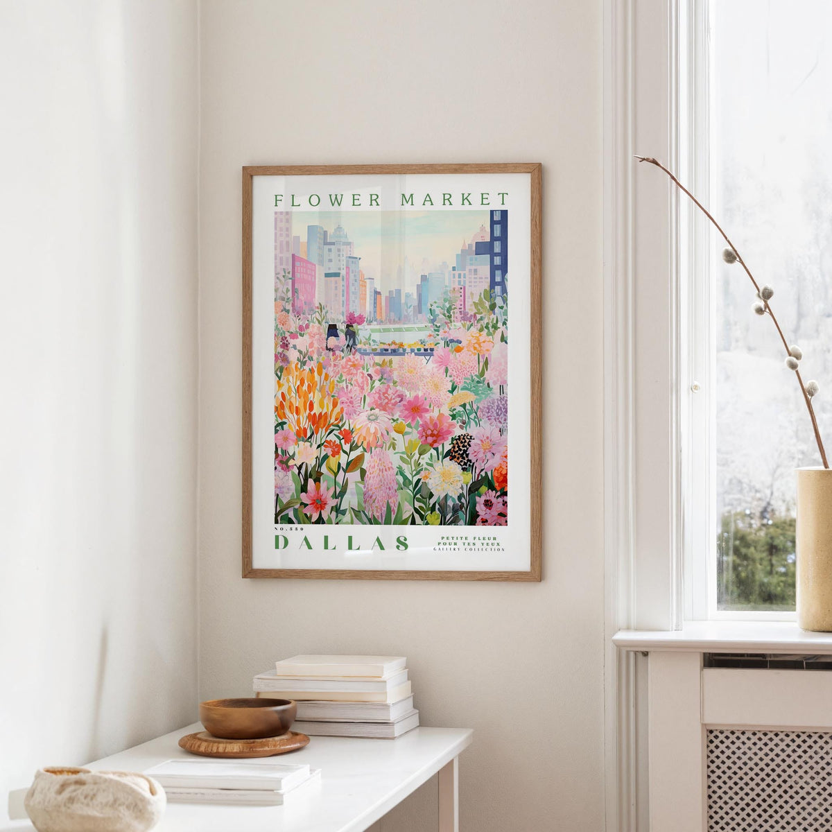 a framed poster of a colorful floral scene hanging on a wall, with a white table and a vase of flowers in the foreground.