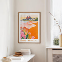 a framed poster of a beach scene with mountains in the background, hanging on a wall in a room with a white table and a vase of flowers.