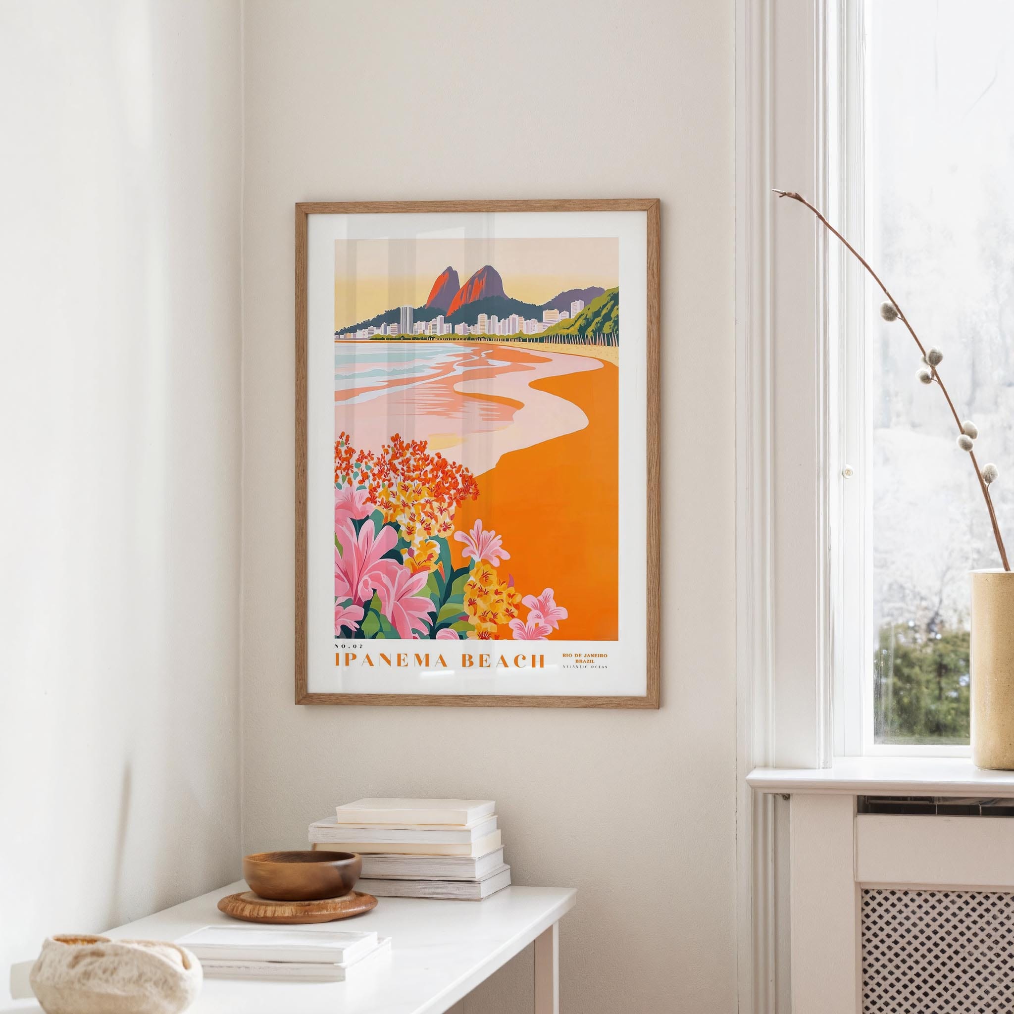 a framed poster of a beach scene with mountains in the background, hanging on a wall in a room with a white table and a vase of flowers.