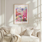 a framed poster of a cityscape with a flower market in the background, hanging on a wall above a white couch with a blanket draped over it.