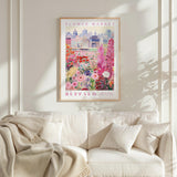 a framed poster of a cityscape with a flower market in the background, hanging on a wall above a white couch with a blanket draped over it.