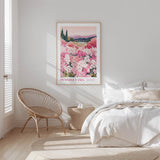 A bedroom with a large bed, a woven chair, and a framed poster of a field of pink flowers hanging on the wall.