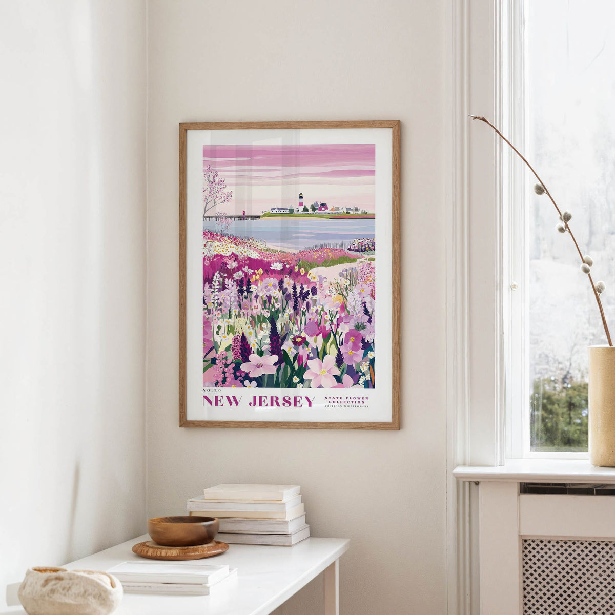 a framed poster of a scenic landscape with a body of water and a small island, hanging on a wall in a room with a window and a table with books and a vase.