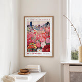 a framed poster of a colorful flower market scene hanging on a wall, with a white table and a vase of flowers in the foreground.