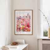a framed poster of a colorful floral scene hanging on a wall, with a white table and a vase of flowers in the foreground.