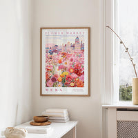 a framed poster of a colorful floral scene hanging on a wall, with a white table and a vase of flowers in the foreground.
