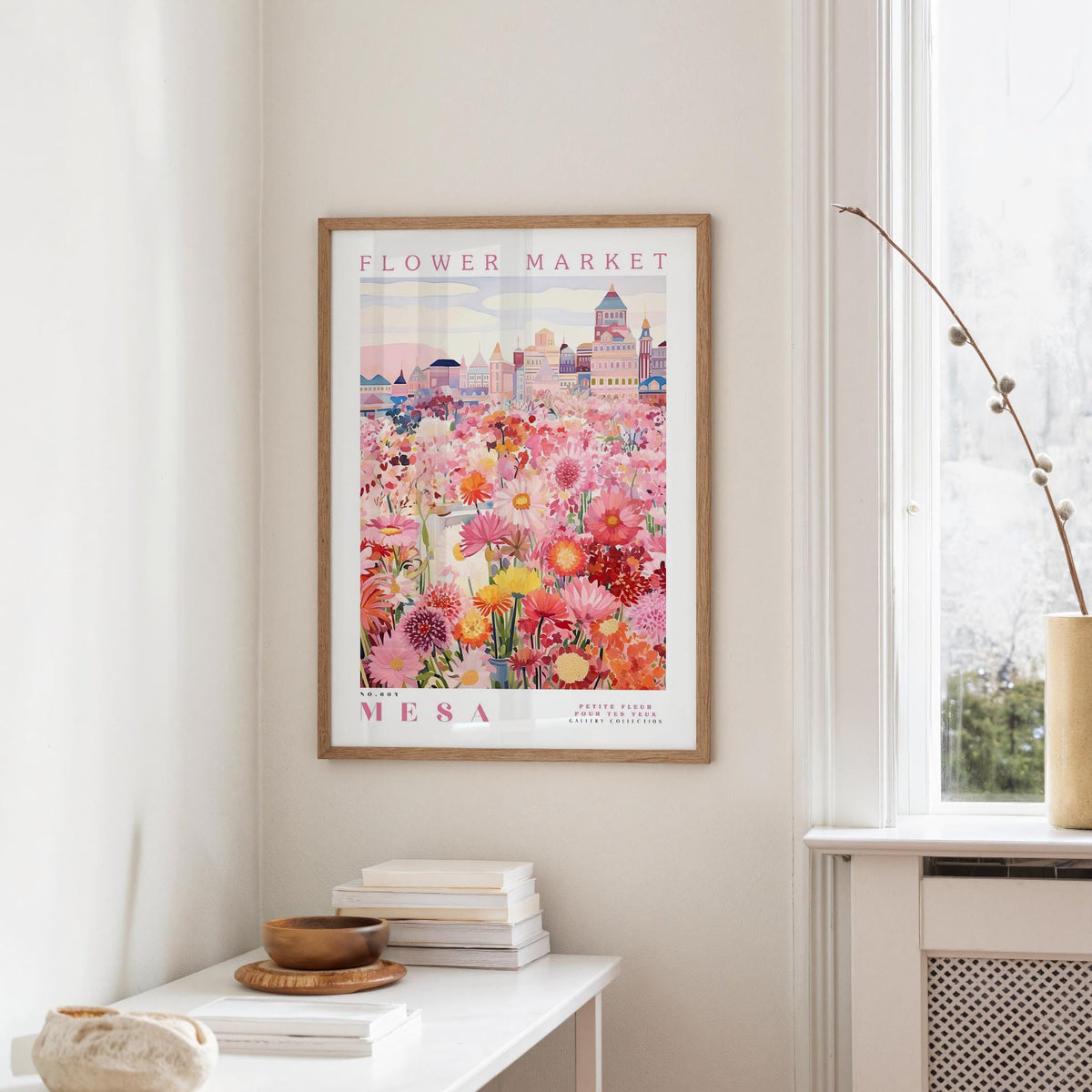 a framed poster of a colorful floral scene hanging on a wall, with a white table and a vase of flowers in the foreground.