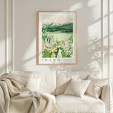 a framed poster of a scenic landscape with mountains, flowers, and a lake, hanging on a wall above a white couch with a blanket draped over it.