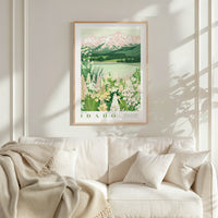a framed poster of a scenic landscape with mountains, flowers, and a lake, hanging on a wall above a white couch with a blanket draped over it.