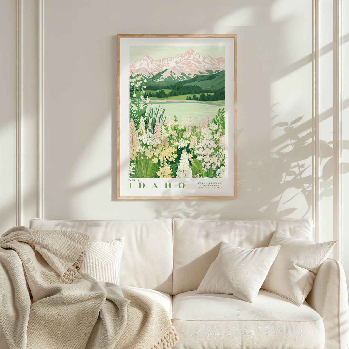 a framed poster of a scenic landscape with mountains, flowers, and a lake, hanging on a wall above a white couch with a blanket draped over it.