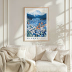 a framed poster of a mountainous landscape with a field of flowers, hanging on a wall above a white couch with a blanket draped over it.