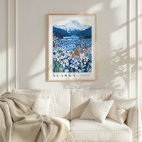 a framed poster of a mountainous landscape with a field of flowers, hanging on a wall above a white couch with a blanket draped over it.