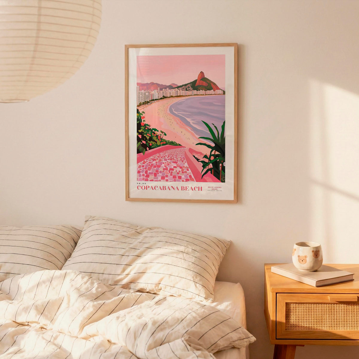 a framed poster of Copacabana Beach hanging on a wall above a bed with striped pillows.