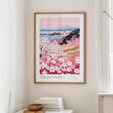 a framed poster of a scenic landscape with a mountain and a field of pink flowers, hanging on a wall in a room with a window and a small table with books and a bowl.