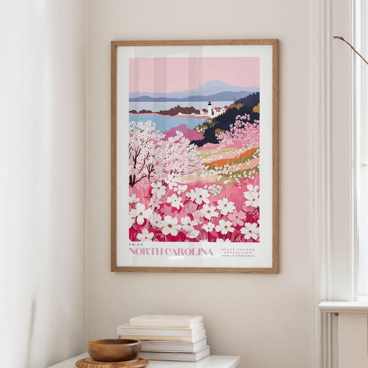 a framed poster of a scenic landscape with a mountain and a field of pink flowers, hanging on a wall in a room with a window and a small table with books and a bowl.