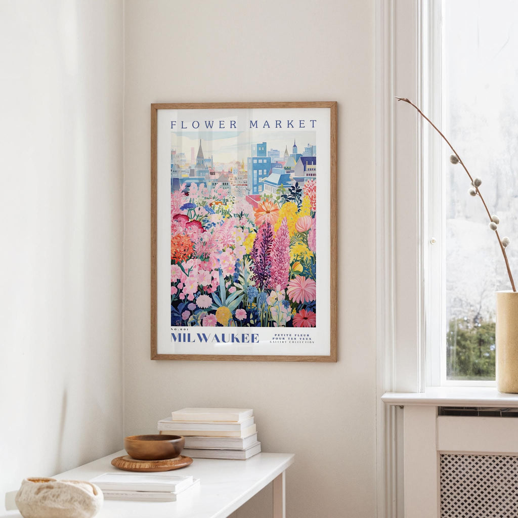 a framed poster of a colorful floral scene hanging on a wall, with a window and a small table with books and a vase in the foreground.