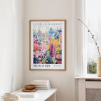 a framed poster of a colorful floral scene hanging on a wall, with a window and a small table with books and a vase in the foreground.