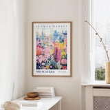 a framed poster of a colorful floral scene hanging on a wall, with a window and a small table with books and a vase in the foreground.
